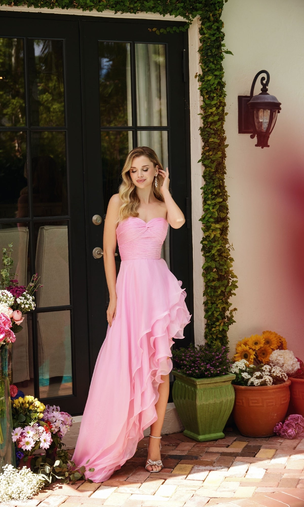 A woman in the Strapless High-Low Prom Dress Ava Presley 40055, featuring a pink asymmetrical ruffle design and flowing skirt, stands outside by glass doors amidst potted plants and colorful flowers.